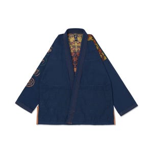 Op de afbeelding: Een donkerblauw vechtsport-gi-jasje met een textuurpaneel aan de onderkant, lange mouwen met cirkelvormige geborduurde emblemen en een levendige, meerkleurig bedrukte binnenvoering zichtbaar bij de kraag en schouder.
