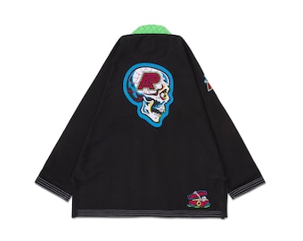 May include: Black martial arts gi with a neon green collar and a large skull graphic on the back. The skull is white, yellow, and blue, with a red letter "R" inside. Additional patches are on the sleeves and lower back.