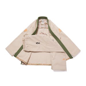 May include: A tan martial arts uniform with a green collar and trim. The uniform includes a jacket and trousers. The jacket has a decorative pattern on the inside and patches on the sleeves. The trousers have a drawstring waist and a small patch.