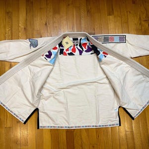 Op de afbeelding: Crèmekleurig vechtsportuniform met een patroon aan de binnenkant met rode, zwarte en blauwe vormen. Het uniform heeft een patch op de mouw en decoratieve randen. De gi ligt op een houten oppervlak.