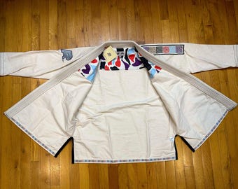 May include: Cream-coloured martial arts uniform with a patterned interior featuring red, black, and blue shapes. The uniform has a patch on the sleeve and decorative trim. The gi is laid out on a wooden surface.