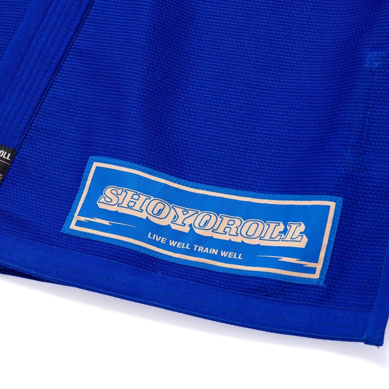 May include: Close-up of a royal blue martial arts uniform. A rectangular patch with the text "SHOYOROLL" in a stylized font is sewn onto the fabric. The patch also includes the text "LIVE WELL TRAIN WELL".