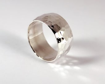 May include: A wide, silver-colored ring with a hammered texture. The ring has a thick band and a shiny, reflective surface. The ring is displayed on a white background, with shadows adding depth.