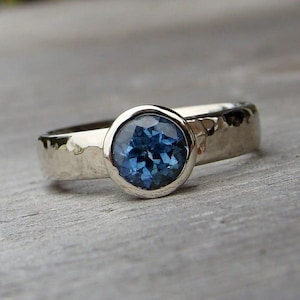 Puede incluir: Un anillo de plata con una piedra preciosa azul oscuro, redonda y facetada, engastada en un bisel. La banda tiene una textura martillada y el anillo se muestra sobre una superficie de madera.