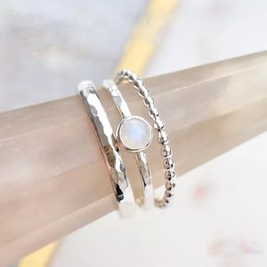 May include: A set of three silver rings. One ring has a hammered texture, another features a small, round, iridescent stone, and the third has a beaded design. The rings are displayed on a light-colored surface.