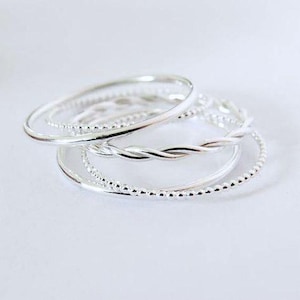 May include: A collection of silver-colored rings in various designs. The rings include a smooth band, a twisted rope design, and a beaded band. The rings are stacked together, creating a layered effect. The jewelry is displayed on a white surface.