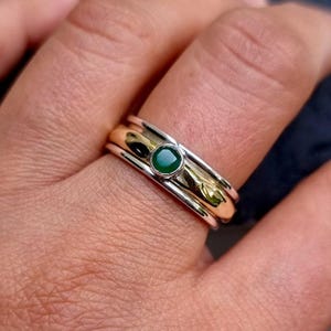 May include: A close-up of a ring with a gold band, a silver band, and a green gemstone. The ring is on a finger. The gemstone is a deep emerald green colour. The ring has a spinning design.