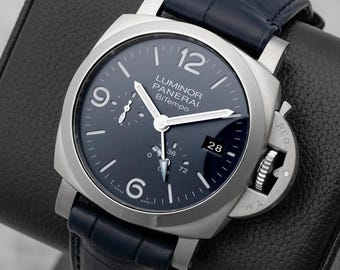 May include: A stainless steel watch with a dark blue face and a matching leather band. The watch face displays the text "LUMINOR PANERAI BiTempo" and the date "28". The watch is displayed on a black leather stand.