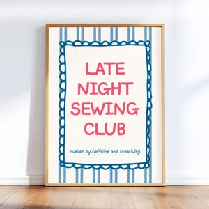 May include: A framed print with the text "LATE NIGHT SEWING CLUB" in pink, surrounded by a blue scalloped border on a cream background. The background has blue vertical stripes. The text "Fuelled by caffeine and creativity" is at the bottom.