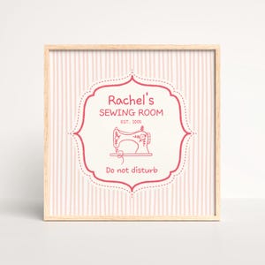 May include: A framed print with a light wood frame. The print has a pink and white striped background and a decorative label in the centre. The label reads "Rachel's SEWING ROOM" with a sewing machine illustration and "Do not disturb."