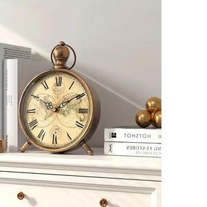 May include: A bronze-coloured, round clock with a world map face and Roman numerals. The clock sits on a white surface, alongside books and gold spheres. The clock's design evokes a vintage aesthetic.