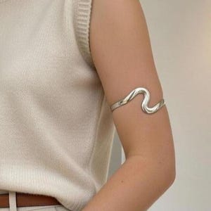 May include: A silver-toned arm cuff with a wavy design. The cuff is worn on the upper arm and has a smooth, polished finish. The jewelry is a modern accessory.