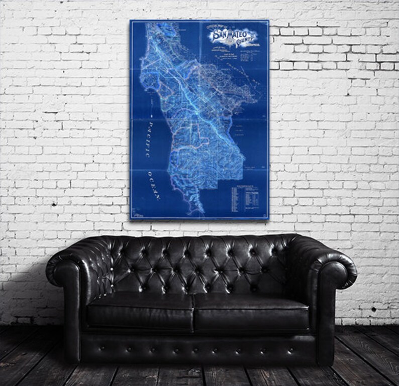 May include: A vintage blue and white map of San Mateo County, California. The map shows the county's roads, rivers, and towns. The map is framed and hanging on a white brick wall.