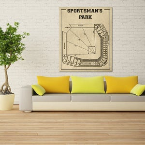 May include: A vintage-style illustration of a baseball field, labeled "Sportsman's Park". The illustration is printed on a beige background and framed with a white border. The illustration is displayed on a white brick wall above a gray couch with yellow throw pillows.