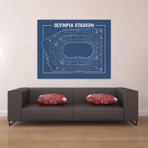 May include: A blueprint style print of Olympia Stadium, a hockey arena in Detroit, Michigan. The print is on a blue background with white lines and text. The text reads "Olympia Stadium" and includes the seating chart for the arena.