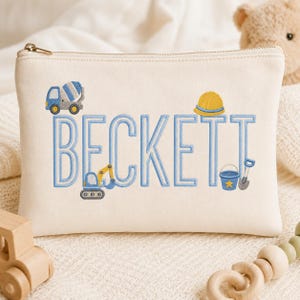 Puede incluir: Un estuche de lona color crema con cremallera que presenta el nombre BECKETT bordado en letras azul claro. El diseño incluye una hormigonera azul y gris, un casco de construcción amarillo, una excavadora azul, un cubo azul y una pala gris.