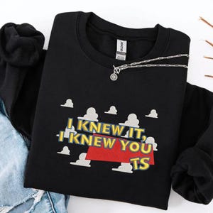 May include: A black long-sleeve sweatshirt featuring a graphic print of white clouds against a blue sky background. The text reads I KNEW IT, I KNEW YOU TS in yellow letters with a red trapezoid shape underneath the text on the front of the garment.