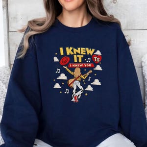 May include: A navy blue crewneck sweatshirt featuring a graphic print of a person with long hair playing a guitar. The text reads I KNEW IT and I KNEW YOU, accompanied by a red hat, a red heart with TS, musical notes, stars, and white clouds.