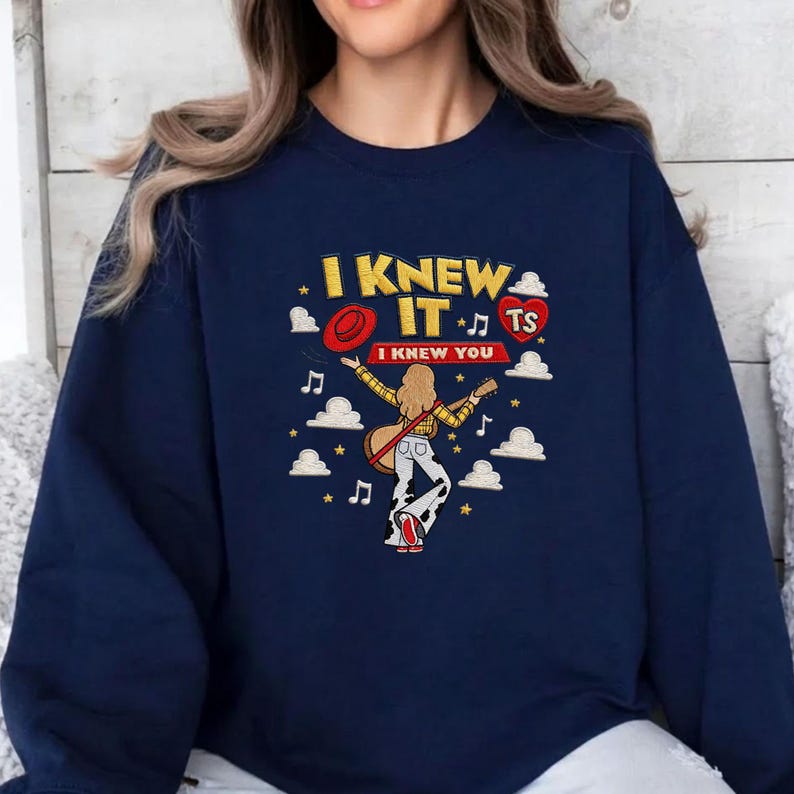 May include: A navy blue crewneck sweatshirt featuring a graphic print of a person with long hair playing a guitar. The text reads I KNEW IT and I KNEW YOU, accompanied by a red hat, a red heart with TS, musical notes, stars, and white clouds.