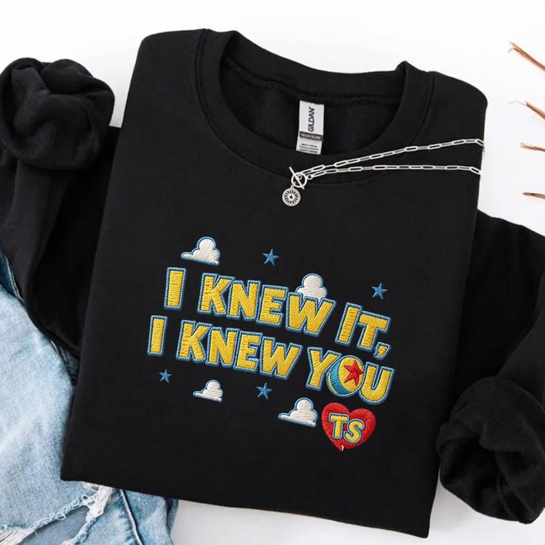 Op de afbeelding: Een zwart sweatshirt met ronde hals en geel borduursel met blauwe omlijning met de tekst "I KNEW IT, I KNEW YOU". Het ontwerp bevat kleine blauwe sterren, witte wolken en een rood hart met "TS" in het geel, plus een zilveren ketting.
