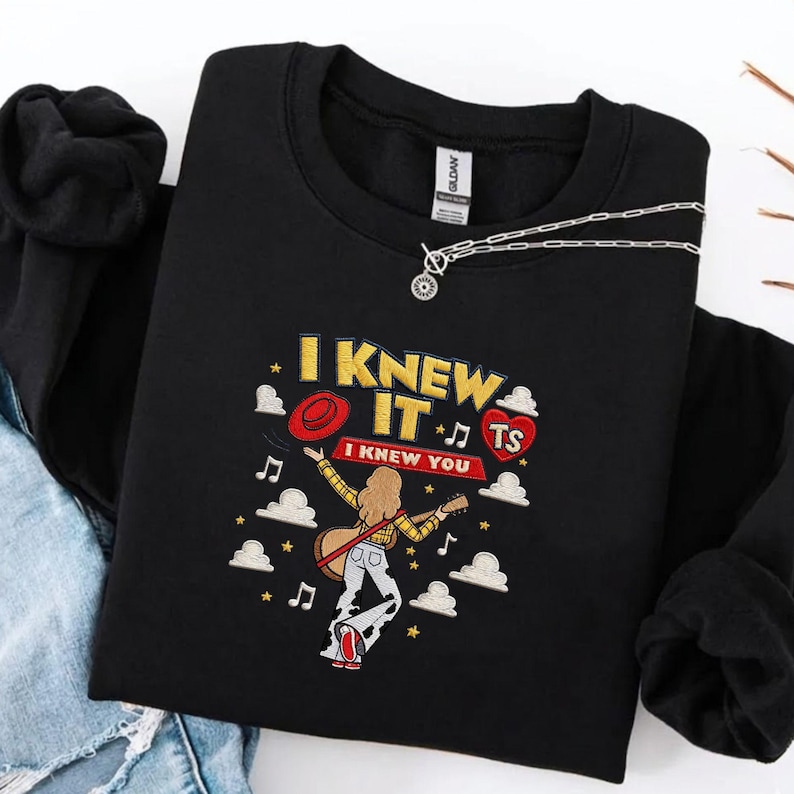 May include: A black crewneck sweatshirt featuring an embroidered graphic of a person with long hair playing a guitar, surrounded by clouds, musical notes, and stars. The text reads I KNEW IT and I KNEW YOU, with a red hat and a red heart labeled TS.
