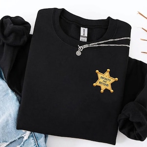 May include: A black long-sleeve sweatshirt featuring a gold embroidered sheriff star badge on the left chest. The badge contains the black text "INFINITY AND BEYOND". A silver-toned chain necklace with a circular pendant rests on the neckline.