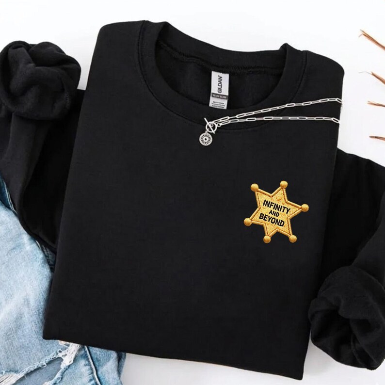 May include: A black long-sleeve sweatshirt featuring a gold embroidered sheriff star badge on the left chest. The badge contains the black text "INFINITY AND BEYOND". A silver-toned chain necklace with a circular pendant rests on the neckline.