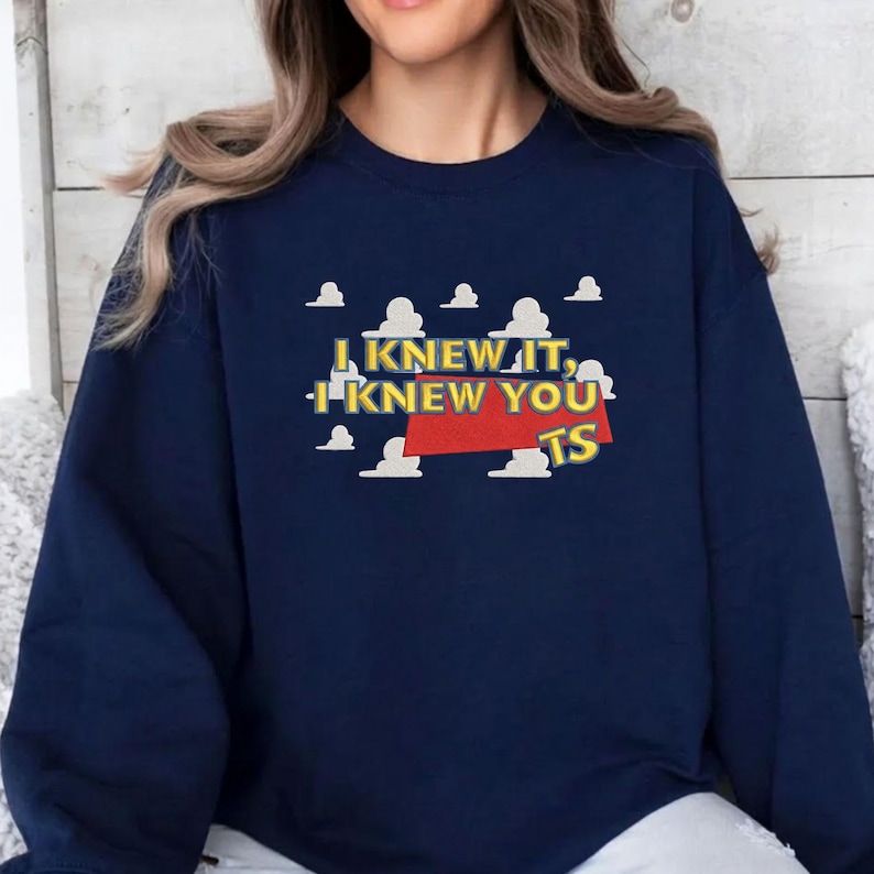 May include: A navy blue crewneck sweatshirt featuring a graphic print of white clouds against a blue sky background. The text "I KNEW IT, I KNEW YOU TS" is printed in yellow letters over a red rectangular shape centered on the chest area.