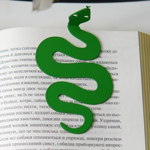 Pode incluir: Um marcador de livros verde em forma de cobra está nas páginas de um livro. O marcador de livros é feito de um material verde sólido e tem um design simples e estilizado. O livro está aberto, com texto visível nas páginas.