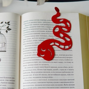 May include: A red snake-shaped bookmark rests on the pages of an open book. The bookmark has a stylized design with cut-out details. The book has text in a foreign language and a drawing of a birdcage.