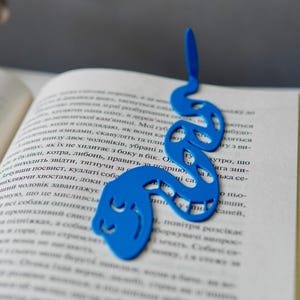 May include: A blue, snake-shaped bookmark rests on the pages of an open book. The bookmark is made of a solid material and features a stylized design with cut-out details. The book's pages are filled with text.