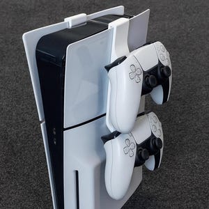 Peut inclure: Une console de jeu blanche et noire avec deux manettes blanches fixées à un support blanc. La console est verticale, avec un design épuré. Les manettes sont blanches avec des accents noirs.