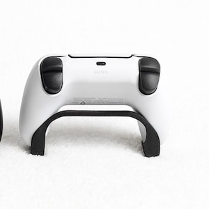 Könnte beinhalten: Zwei Gaming-Controller, einer schwarz mit gelben Akzenten und der andere weiß, werden nebeneinander auf einer weißen, strukturierten Oberfläche gezeigt. Der schwarze Controller hat ein schlankes Design mit mehreren Tasten. Der weiße Controller hat ein geschwungenes Design.