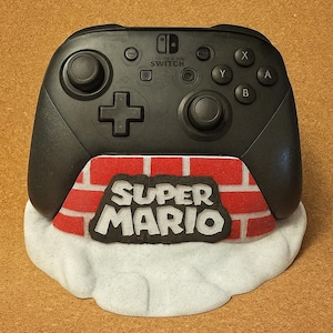 Op de afbeelding: Zwarte Nintendo Switch Pro Controller met het Super Mario-logo op een bakstenen muurontwerp. De controller staat op een witte wolk-vormige basis. De controller heeft een standaard knoppenindeling.