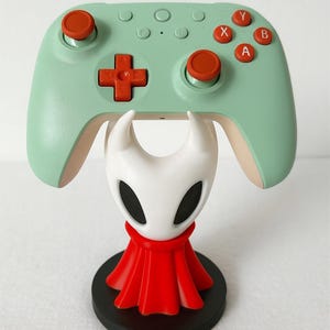 Può includere: Un controller per videogiochi verde menta con pulsanti arancioni e un pad direzionale rosso poggia su un supporto per personaggi bianco e rosso. Il supporto ha una base nera. Il controller ha le lettere X, Y, A e B.