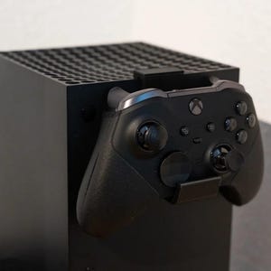 May include: A black Xbox Series X console with a black controller attached to a black stand. The controller has a silver accent and multiple buttons and joysticks. The console has a perforated top for ventilation.