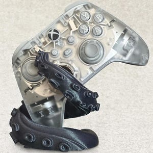 May include: A clear Xbox controller with a dark gray octopus-shaped grip. The controller's internal components are visible. The octopus grip wraps around the controller, adding a unique aesthetic to the gaming accessory.