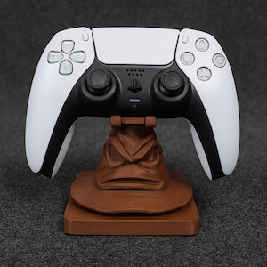 Puede incluir: Un mando blanco y negro de PlayStation 5 descansa sobre un soporte marrón con forma de Sombrero Seleccionador. El mando tiene un diseño elegante con detalles en negro. El soporte es una réplica detallada del Sombrero Seleccionador.