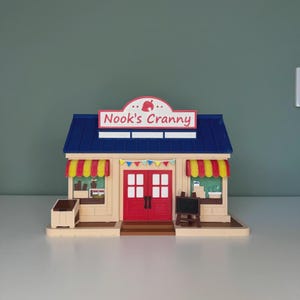 Op de afbeelding: Een miniatuur houten replica van Nook's Cranny, een winkel uit de Animal Crossing-videogame. Het gebouw heeft een blauw dak, rode deuren en luifels met gele en rode strepen. Het bord leest "Nook's Cranny".