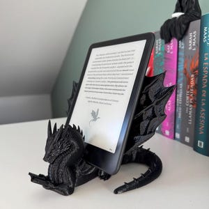 Può includere: Un supporto per e-reader a forma di drago nero che tiene un e-reader con testo. Il supporto presenta squame e ali dettagliate. Sullo sfondo sono visibili libri con copertine colorate.