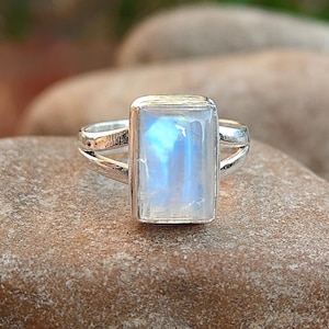 May include: A silver ring with a rectangular, iridescent moonstone. The moonstone has a blue and white glow. The ring is set on a brown stone, with a blurred green background.