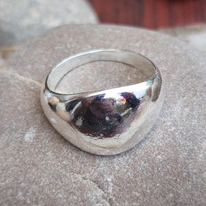 Può includere: Un anello in argento lucido con un design arrotondato e liscio. L'anello è appoggiato su una pietra grigia, che riflette la luce. La fascia dell'anello è larga circa 0,6 cm. Lo stile generale è minimalista e moderno.