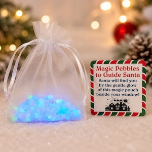 May include: A sheer white organza drawstring pouch filled with small, glowing blue pebbles. Beside the pouch is a square card with a red and green striped border, featuring the text: Magic Pebbles to Guide Santa. Santa will find you by the gentle glow of this magic pouch beside your window! with a silhouette of a sleigh and houses.