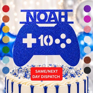 May include: A blue glitter cake topper shaped like a games controller, with the name "NOAH" and the number "10" in white. The topper sits on a white cake with blue icing.