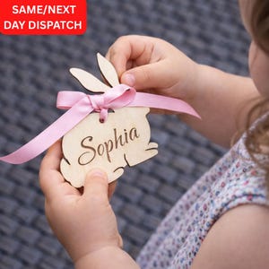 May include: A wooden bunny-shaped name tag with the name "Sophia" engraved. It's tied with a pink ribbon and held by a person. The text "SAME/NEXT DAY DISPATCH" is in a red box in the upper left corner.