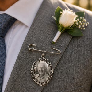 May include: A silver-toned oval photo charm pin featuring an ornate filigree border and a black and white portrait of an older person. The charm is attached to a large safety pin and is pinned to the lapel of a grey textured suit jacket.