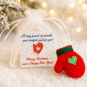 May include: A white sheer organza drawstring pouch with a square tag featuring a red mitten graphic and the text A tiny parcel of warmth and comfort, just for you! Merry Christmas and a Happy New Year! A small red knitted mitten with a green cuff and heart.
