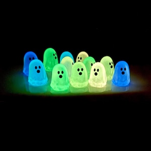 May include: A collection of small, translucent ghost figurines that glow in the dark. Each ghost features a rounded shape with two black oval eyes and a small black circular mouth. The figures emit blue, green, and yellow light against a black background.