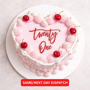 May include: A heart-shaped pink cake decorated with white frosting, pink frosting swirls, and red cherries. The words "twenty one" are written in red script on the cake. Gold sprinkles are also visible. The cake sits on a white plate.