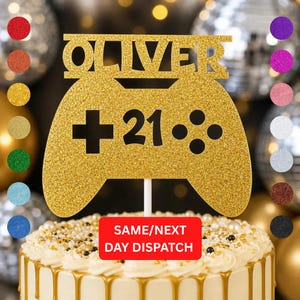 May include: A gold glitter cake topper shaped like a games controller, with the name "OLIVER" and the number "21". The topper sits atop a white cake with gold icing. A red rectangle at the bottom reads "SAME/NEXT DAY DISPATCH".